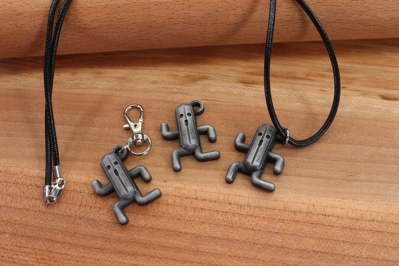 May include: Three silver-colored pendants shaped like a character from a video game, hanging on black cords. One pendant is attached to a silver key chain.