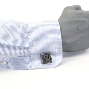 May include: A pair of cufflinks with a silver finish. The cufflinks feature a square design with a stylized symbol inside. The symbol is a circle with a line through it, and a smaller circle inside the larger circle.