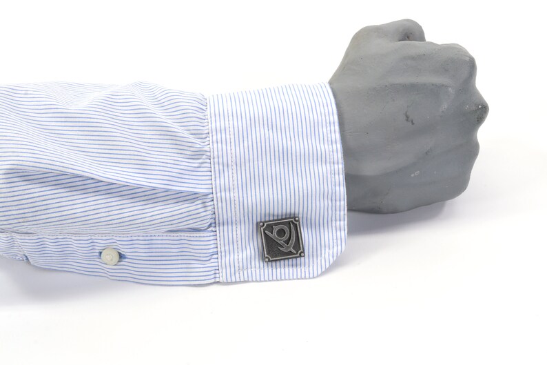May include: A pair of cufflinks with a silver finish. The cufflinks feature a square design with a stylized symbol inside. The symbol is a circle with a line through it, and a smaller circle inside the larger circle.