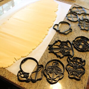 May include: A sheet of rolled out cookie dough on a white parchment paper surface.  A variety of black plastic cookie cutters are arranged on the surface, including characters from a popular video game.