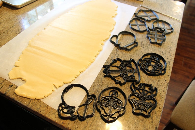 May include: A sheet of rolled out cookie dough on a white parchment paper surface.  A variety of black plastic cookie cutters are arranged on the surface, including characters from a popular video game.