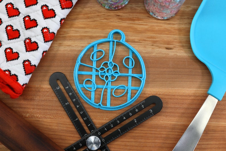 May include: A blue plastic cookie cutter shaped like a circle with a decorative design inside. The cutter is on a wooden surface with other baking tools.