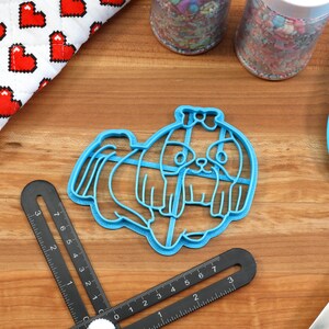 May include: Blue plastic cookie cutter shaped like a Shih Tzu dog. The cutter is on a wooden surface with a ruler and a spatula.