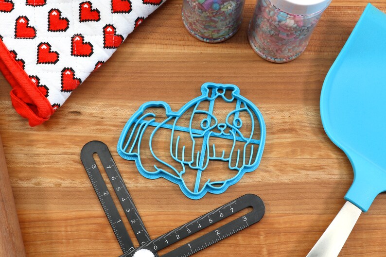 May include: Blue plastic cookie cutter shaped like a Shih Tzu dog. The cutter is on a wooden surface with a ruler and a spatula.