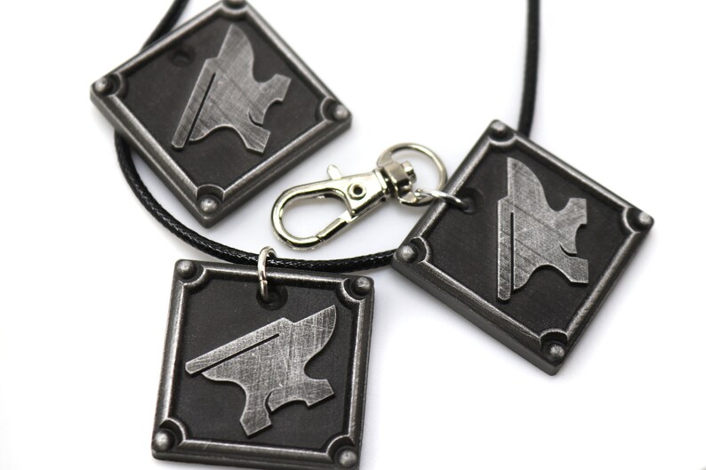 May include: Three silver metal pendants with a black background and an anvil design. The pendants are on a black cord and one is attached to a silver metal key chain.