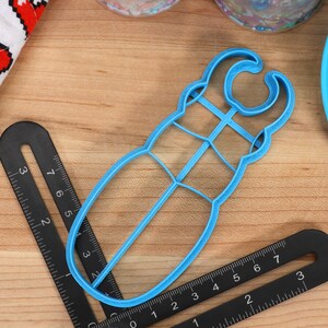 May include: A blue plastic cookie cutter shaped like a beetle. The cutter has a handle on the top and a detailed design.