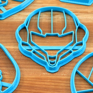 May include: A set of four blue plastic cookie cutters, shaped like a helmet, a flower, a circle, and a rectangle. The cutters are on a wooden surface.