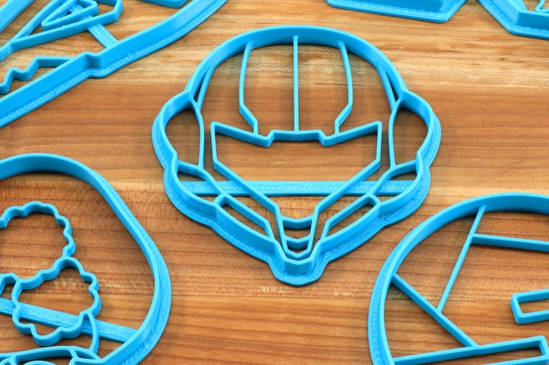 May include: A set of four blue plastic cookie cutters, shaped like a helmet, a flower, a circle, and a rectangle. The cutters are on a wooden surface.