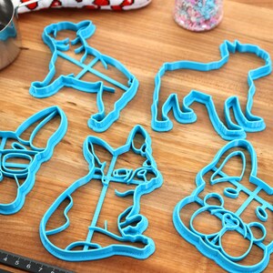 Puede incluir: Un conjunto de cinco cortadores de galletas de pl&aacute;stico azul con forma de diferentes razas de perros. Los cortadores incluyen un bulldog franc&eacute;s con gafas, un bulldog franc&eacute;s sentado, un bulldog franc&eacute;s de pie, un perro con orejas ca&iacute;das y un perro con un hocico largo.