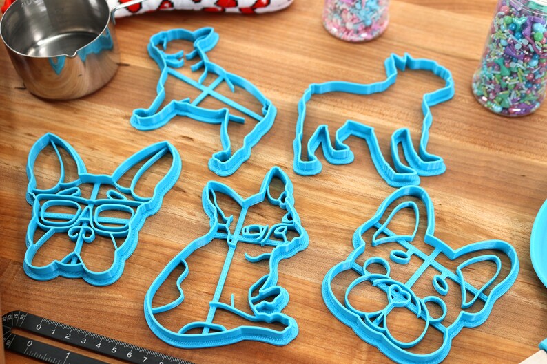 Puede incluir: Un conjunto de cinco cortadores de galletas de pl&aacute;stico azul con forma de diferentes razas de perros. Los cortadores incluyen un bulldog franc&eacute;s con gafas, un bulldog franc&eacute;s sentado, un bulldog franc&eacute;s de pie, un perro con orejas ca&iacute;das y un perro con un hocico largo.