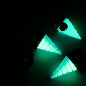 May include: Four glowing teal drill-shaped pendants against a dark background. The pendants have a black top and a spiraled, cone-shaped drill bit that emits a bright teal light. The image is taken from a high angle.