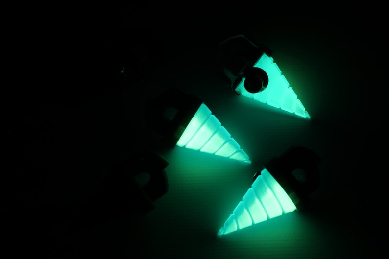 May include: Four glowing teal drill-shaped pendants against a dark background. The pendants have a black top and a spiraled, cone-shaped drill bit that emits a bright teal light. The image is taken from a high angle.