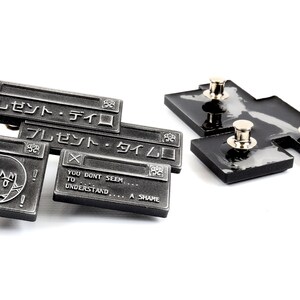 Op de afbeelding: Een set van vier zwarte metalen pinnen met een gepixelde figuur en tekst in het Japans die "Present - Time" luidt. De pinnen zijn ontworpen om eruit te zien als een computerfoutmelding.