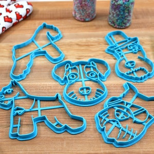 May include: Five blue plastic cookie cutters shaped like different schnauzer dogs. The cutters are on a wooden surface.