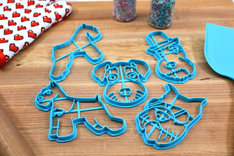 May include: Five blue plastic cookie cutters shaped like different schnauzer dogs. The cutters are on a wooden surface.