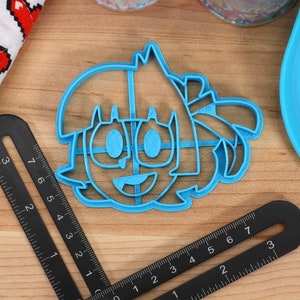 May include: A blue plastic cookie cutter shaped like a cartoon character with a smiling face, large eyes, and hair.