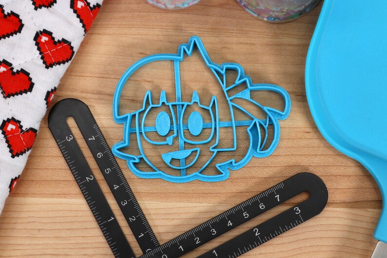 May include: A blue plastic cookie cutter shaped like a cartoon character with a smiling face, large eyes, and hair.