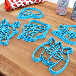 Peut inclure: Ensemble de d&eacute;coupoirs &agrave; biscuits en plastique bleu repr&eacute;sentant des personnages de dessins anim&eacute;s, dispos&eacute;s sur une surface en bois. Des vermicelles dans des r&eacute;cipients en plastique transparent et un tissu &agrave; motifs rouges et blancs sont &eacute;galement visibles.