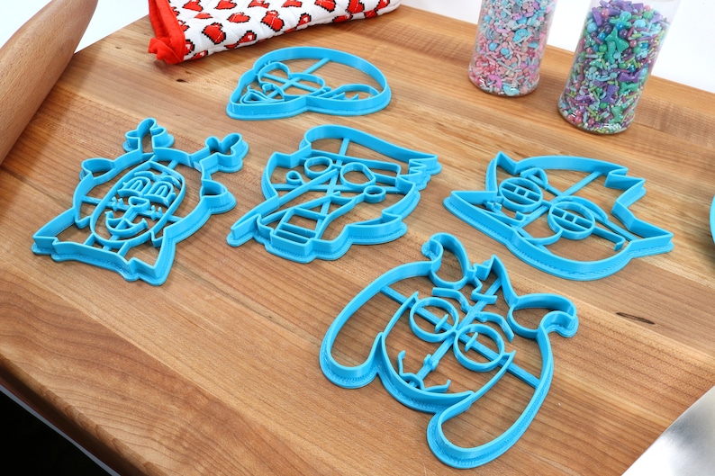 Peut inclure: Ensemble de d&eacute;coupoirs &agrave; biscuits en plastique bleu repr&eacute;sentant des personnages de dessins anim&eacute;s, dispos&eacute;s sur une surface en bois. Des vermicelles dans des r&eacute;cipients en plastique transparent et un tissu &agrave; motifs rouges et blancs sont &eacute;galement visibles.