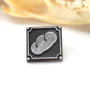 May include: A silver and black metal pin with a stylized image of a loaf of bread.