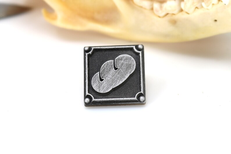 May include: A silver and black metal pin with a stylized image of a loaf of bread.