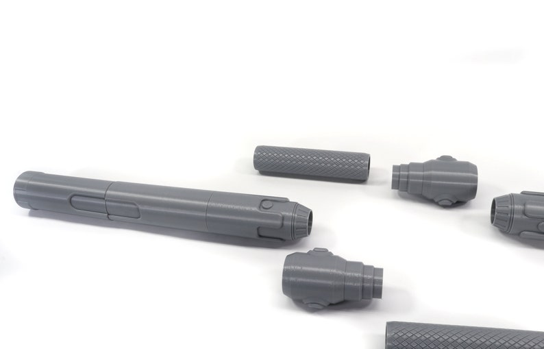 K&ouml;nnte beinhalten: Ein zerlegtes, graues 3D-gedrucktes Modell einer futuristischen Waffe. Die Waffe besteht aus f&uuml;nf Teilen: einem langen Lauf, zwei Endkappen und zwei k&uuml;rzeren Teilen.