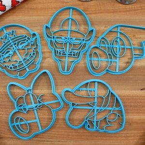 May include: Five blue plastic cookie cutters shaped like cartoon characters. The characters include a mushroom, a turtle, a princess, a ghost, and a man with a mustache.