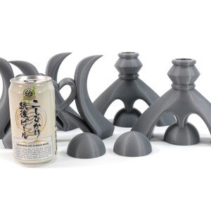 May include: Four gray ceramic candle holders with a unique, abstract design. The holders are shaped like a stylized bird with a rounded base and a tall, curved neck. A can of Japanese beer is in the center of the image.