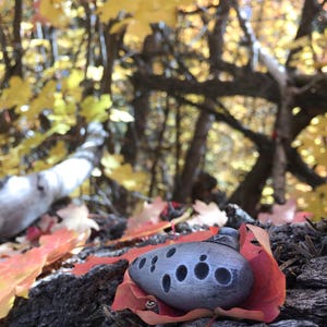 Puede incluir: Una ocarina plateada con un dise&ntilde;o triangular en la parte superior, descansando sobre un lecho de hojas de oto&ntilde;o rojas y amarillas.