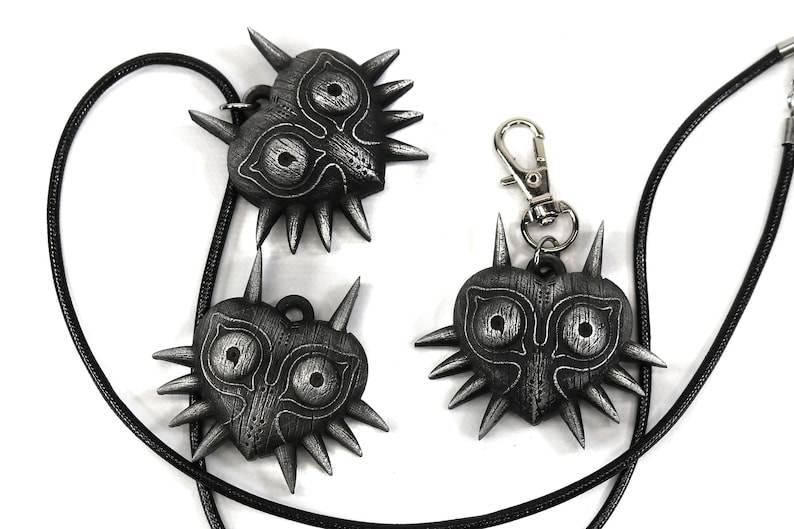 May include: Three silver Majora's Mask pendants, two on black cords and one on a silver chain with a clasp. The masks are heart-shaped with a spiky design and a detailed face.