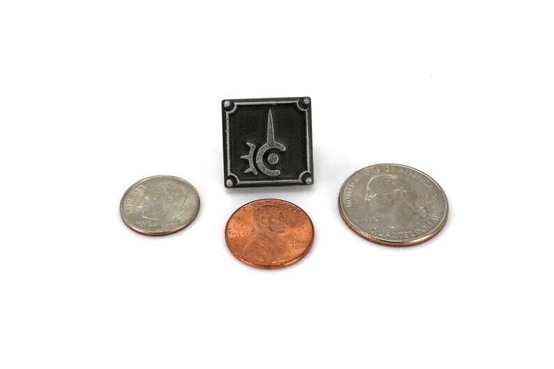 May include: A black and silver metal pin with a stylized sword and circle design. The pin is about the size of a US quarter.
