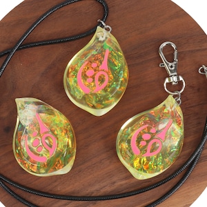 May include: Three teardrop-shaped pendants with a pink symbol inside. The pendants are made of clear resin with iridescent glitter. One pendant is on a black cord necklace, and the other two are on a black cord keychain.