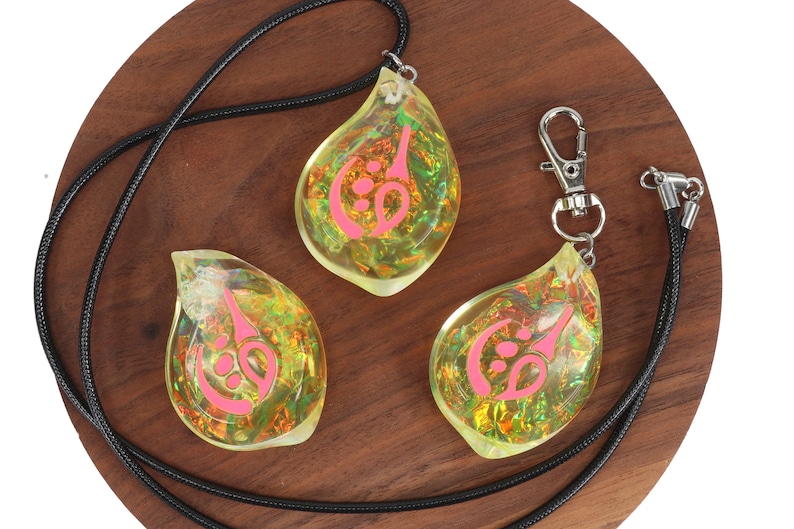 May include: Three teardrop-shaped pendants with a pink symbol inside. The pendants are made of clear resin with iridescent glitter. One pendant is on a black cord necklace, and the other two are on a black cord keychain.