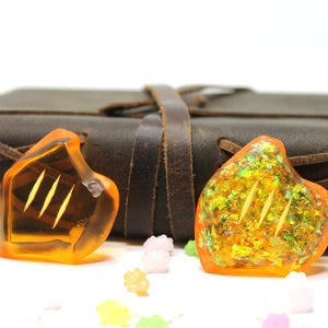 May include: Two orange glass heart-shaped charms with gold glitter and a three-line design. The charms are sitting on a white surface in front of a brown leather-bound journal.