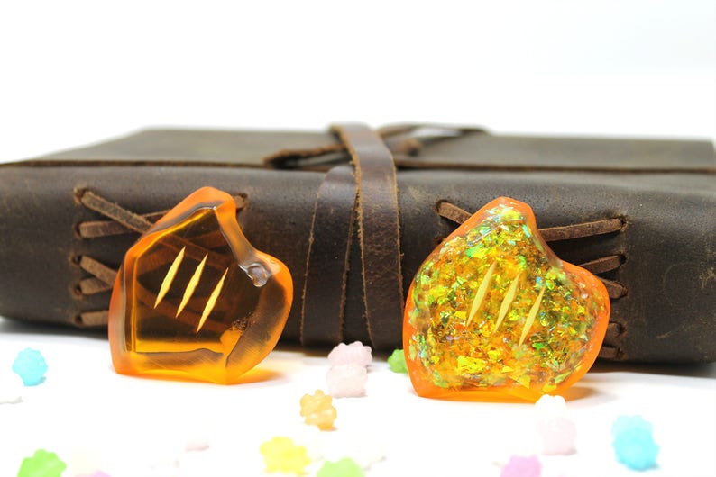 May include: Two orange glass heart-shaped charms with gold glitter and a three-line design. The charms are sitting on a white surface in front of a brown leather-bound journal.