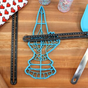 May include: A blue plastic cookie cutter shaped like a hand holding a heart. The cutter is on a wooden surface with a black ruler, a rolling pin, a blue spatula, and a red and white oven mitt.