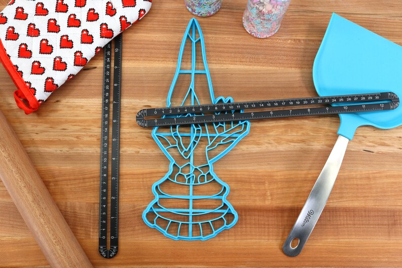 May include: A blue plastic cookie cutter shaped like a hand holding a heart. The cutter is on a wooden surface with a black ruler, a rolling pin, a blue spatula, and a red and white oven mitt.