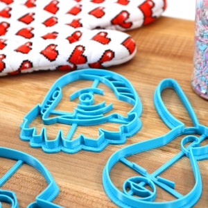 Pu&ograve; includere: Quattro tagliabiscotti in plastica blu a forma di navicella spaziale, coniglio, stella e personaggio con una grande testa. I tagliabiscotti sono su una superficie di legno.