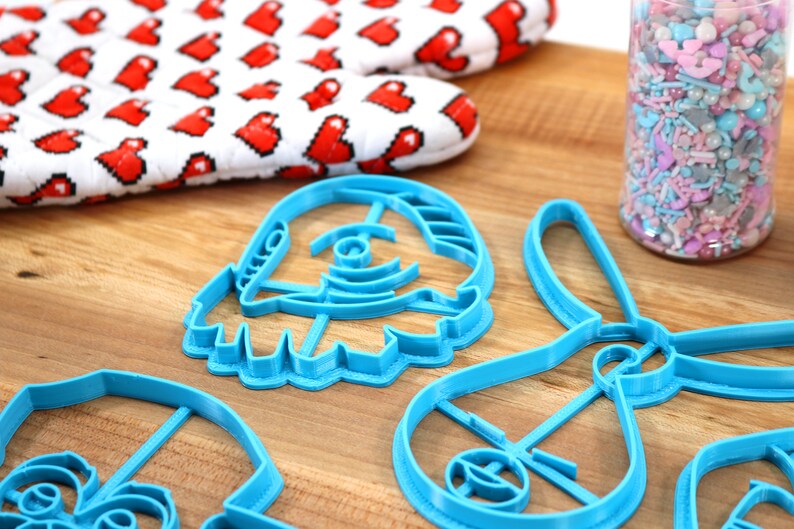 Pu&ograve; includere: Quattro tagliabiscotti in plastica blu a forma di navicella spaziale, coniglio, stella e personaggio con una grande testa. I tagliabiscotti sono su una superficie di legno.