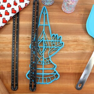May include: A blue plastic cookie cutter shaped like a hand holding a knife. The cutter is on a wooden surface with a rolling pin, measuring tapes, and a blue spatula.