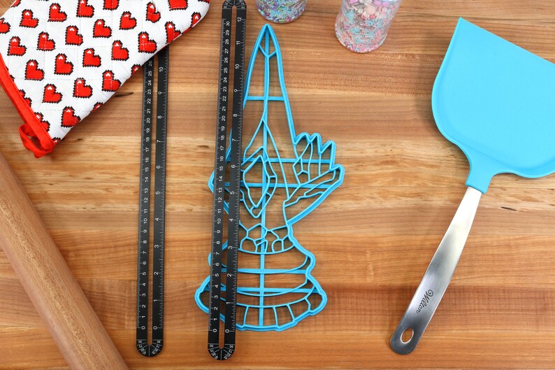 May include: A blue plastic cookie cutter shaped like a hand holding a knife. The cutter is on a wooden surface with a rolling pin, measuring tapes, and a blue spatula.