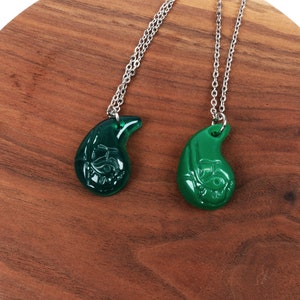 May include: Two green teardrop-shaped necklaces with silver chains. The pendants have a detailed design and are on a wooden surface.