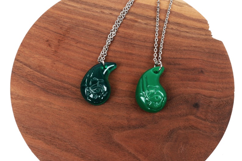 May include: Two green teardrop-shaped necklaces with silver chains. The pendants have a detailed design and are on a wooden surface.