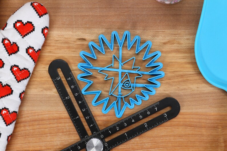 May include: A blue plastic cookie cutter shaped like a cartoon character with a starburst design around the edge. The cookie cutter is on a wooden surface with a black ruler.