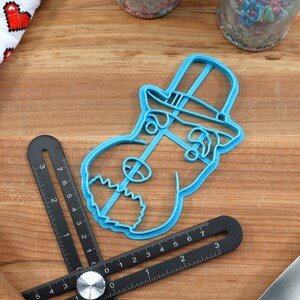May include: Blue plastic cookie cutter shaped like a dog wearing a top hat. The cookie cutter is on a wooden surface.
