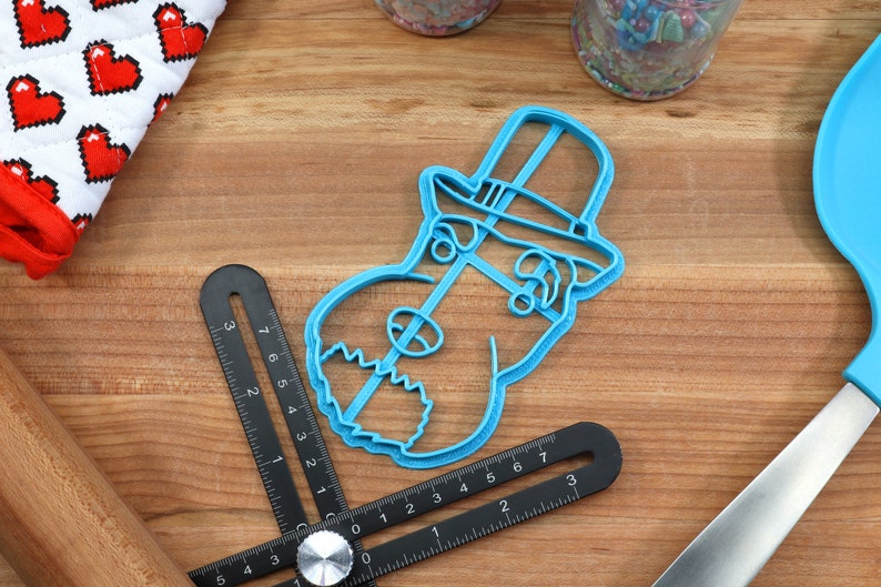 May include: Blue plastic cookie cutter shaped like a dog wearing a top hat. The cookie cutter is on a wooden surface.