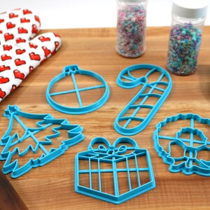 Op de afbeelding: Een set van zes blauwe plastic kerstkoekjesvormen in de vorm van een kerstboom, een snoepstok, een cadeau, een kerstbal, een krans en een ster. De vormen liggen op een houten oppervlak.