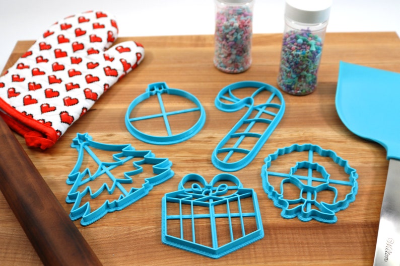 Op de afbeelding: Een set van zes blauwe plastic kerstkoekjesvormen in de vorm van een kerstboom, een snoepstok, een cadeau, een kerstbal, een krans en een ster. De vormen liggen op een houten oppervlak.