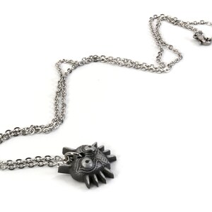 May include: A silver chain necklace with a pendant shaped like the Majora's Mask from the Legend of Zelda video game series. The mask is a dark gray color with intricate details.