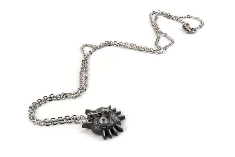 May include: A silver chain necklace with a pendant shaped like the Majora's Mask from the Legend of Zelda video game series. The mask is a dark gray color with intricate details.